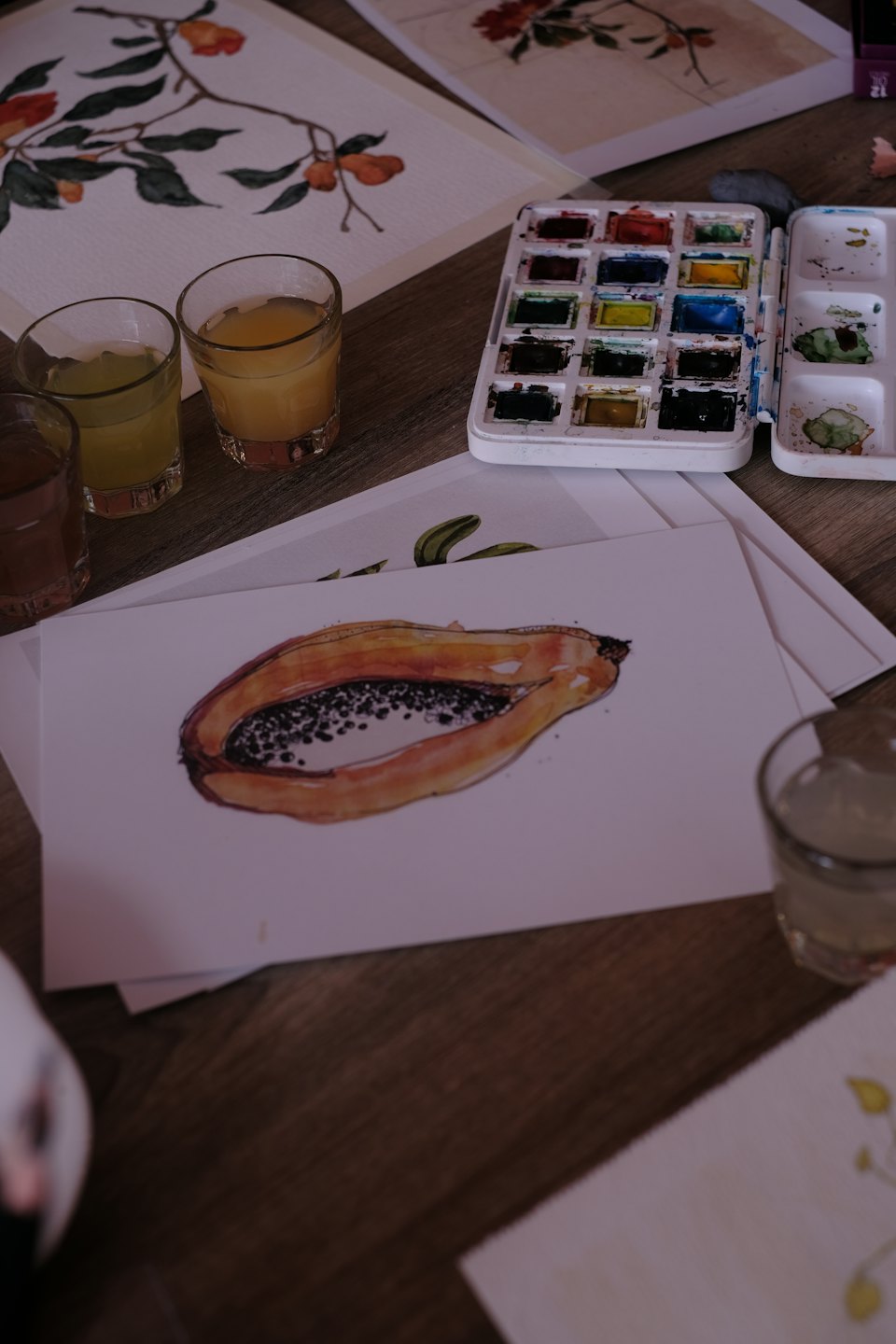 Watercolor supplies and painted illustrations rest on a wooden table.
